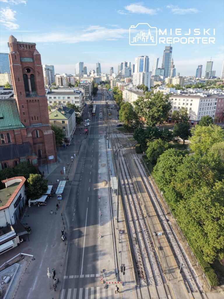Widok na ruchliwą ulicę z torami tramwajowymi, nowoczesne wieżowce w tle, zieleń drzew po bokach.