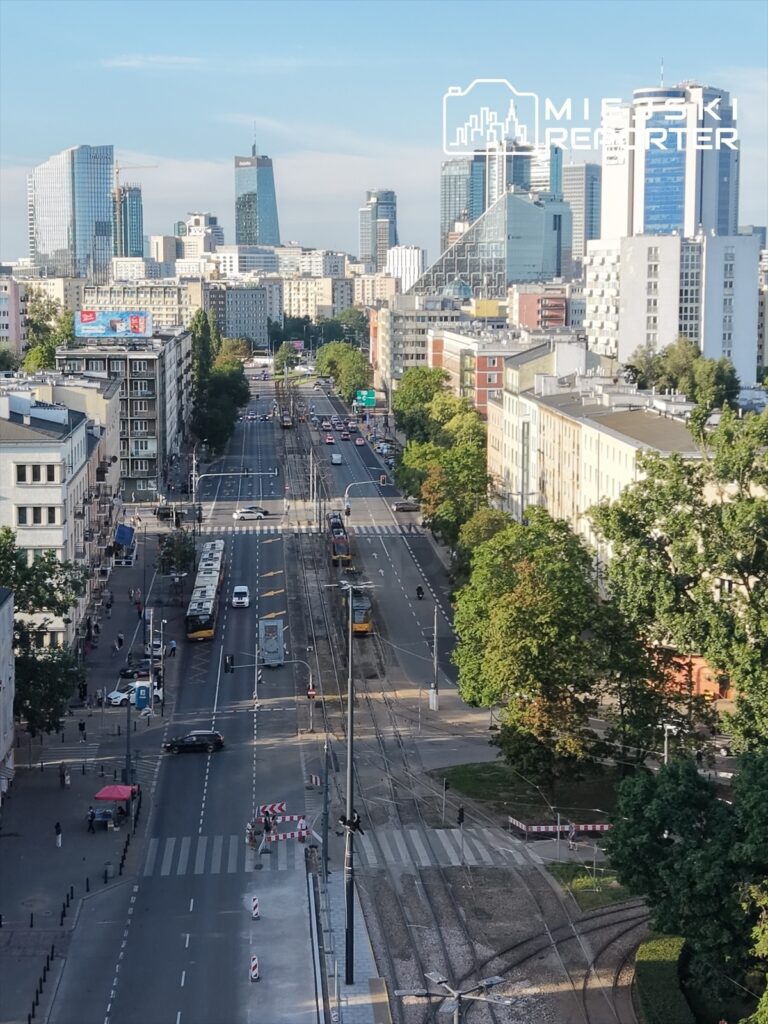 Widok na ruchliwą ulicę w mieście z nowoczesnymi wieżowcami w tle, tramwaje i samochody poruszają się po jezdni.