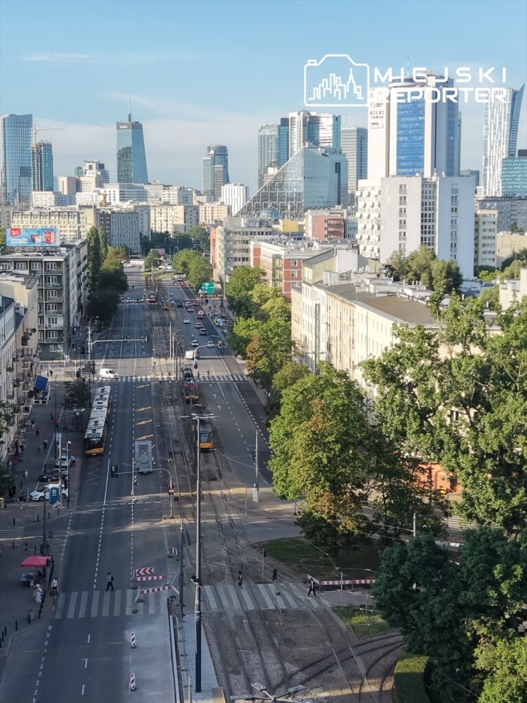 Nowoczesne miasto z wysokimi wieżowcami, ruchliwą ulicą i tramwajami, otoczone zielenią drzew.