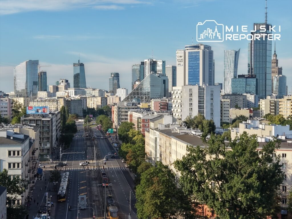 Widok na nowoczesną panoramę miasta z wysokimi wieżowcami, ulicą i tramwajami w tle.