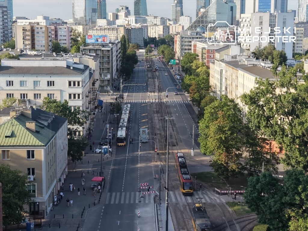 Widok na ruchliwą ulicę z tramwajami i nowoczesnymi wieżowcami w tle, w otoczeniu zieleni i zabudowy miejskiej.