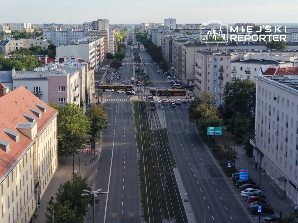 Ruch uliczny na szerokiej drodze z tramwajem i samochodami w miejskim krajobrazie, w tle bloki mieszkalne.
