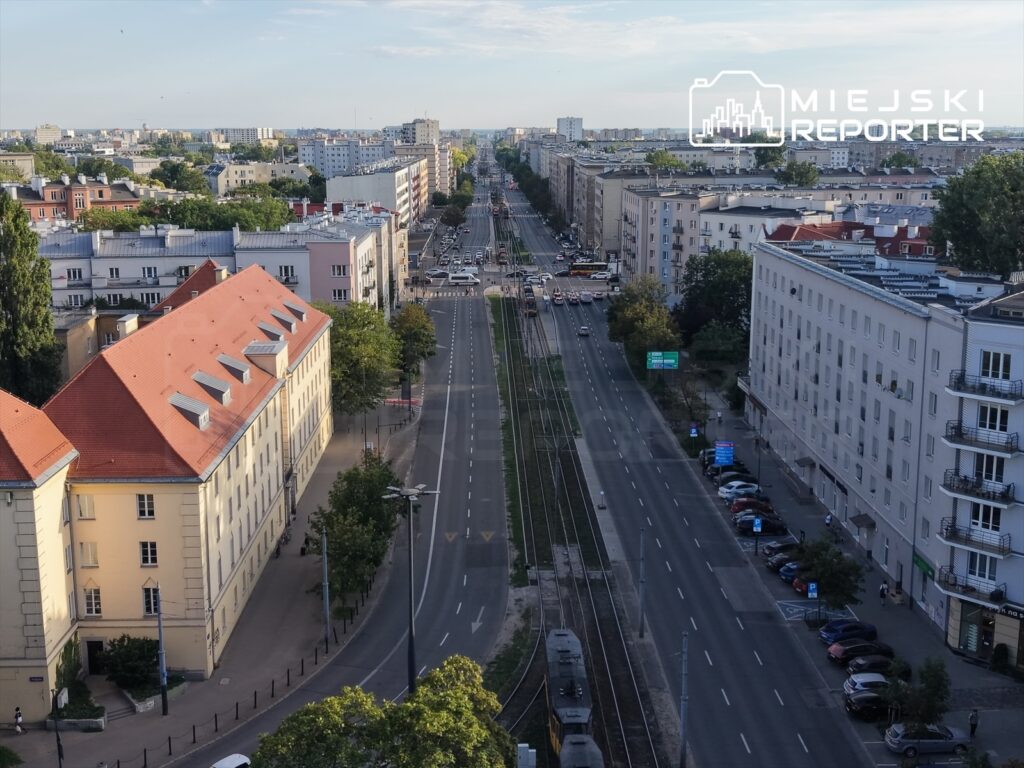 Widok na ruchliwą ulicę z torami tramwajowymi, otoczoną nowoczesnymi budynkami i drzewami w zieleni.