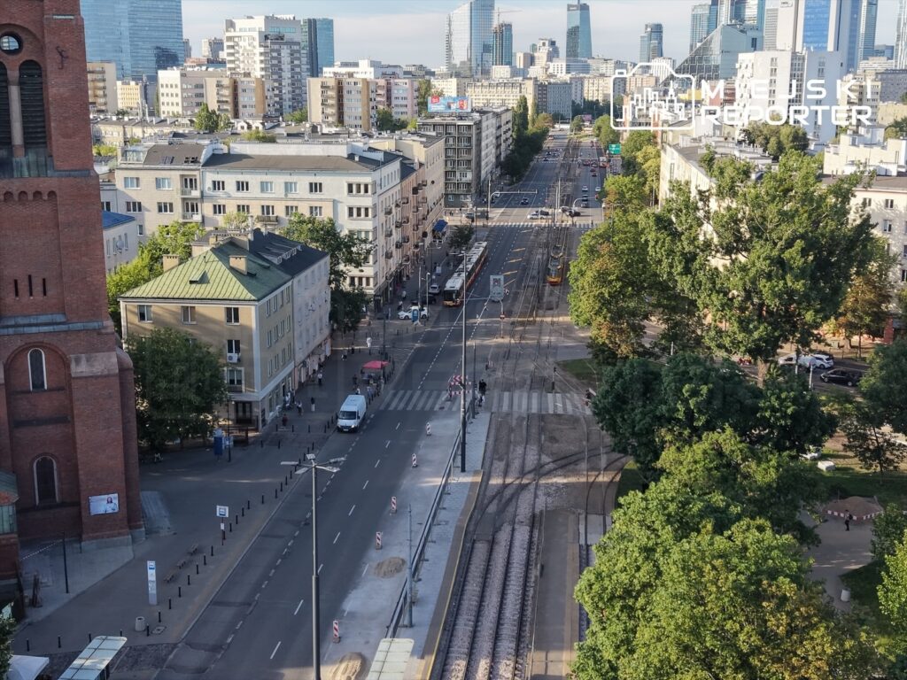 Widok na ruchliwą ulicę z tramwajami, otoczoną nowoczesnymi budynkami i zielenią drzew, w tle wieżowce.