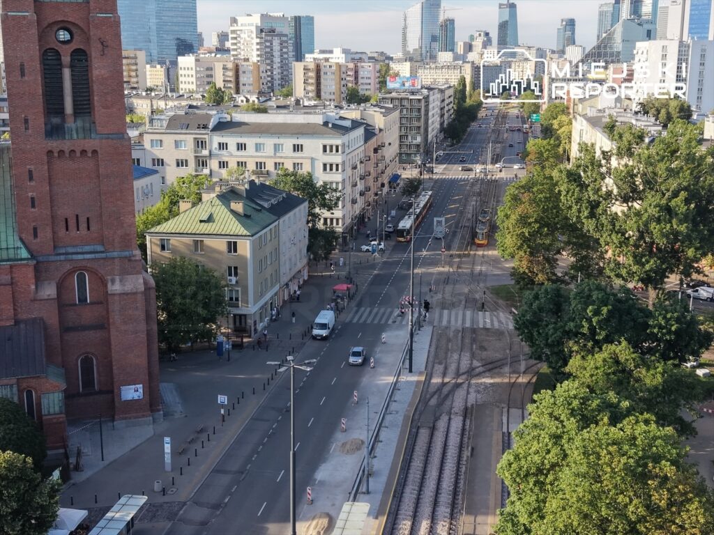 Wysoki, ceglany kościół z wieżą w tle, nowoczesne budynki oraz tramwaje na ulicy z drzewami po bokach.