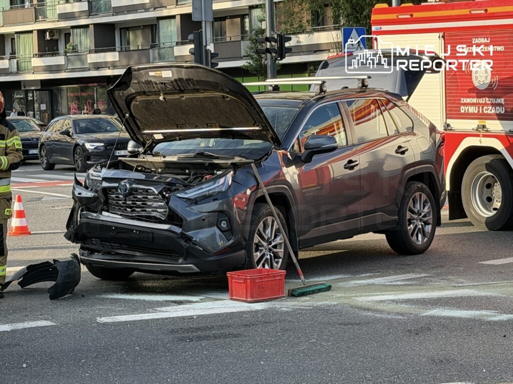 Strażak w mundurze stoi obok uszkodzonego samochodu osobowego z otwartą maską na ulicy, a obok parkowany jest wóz strażacki.