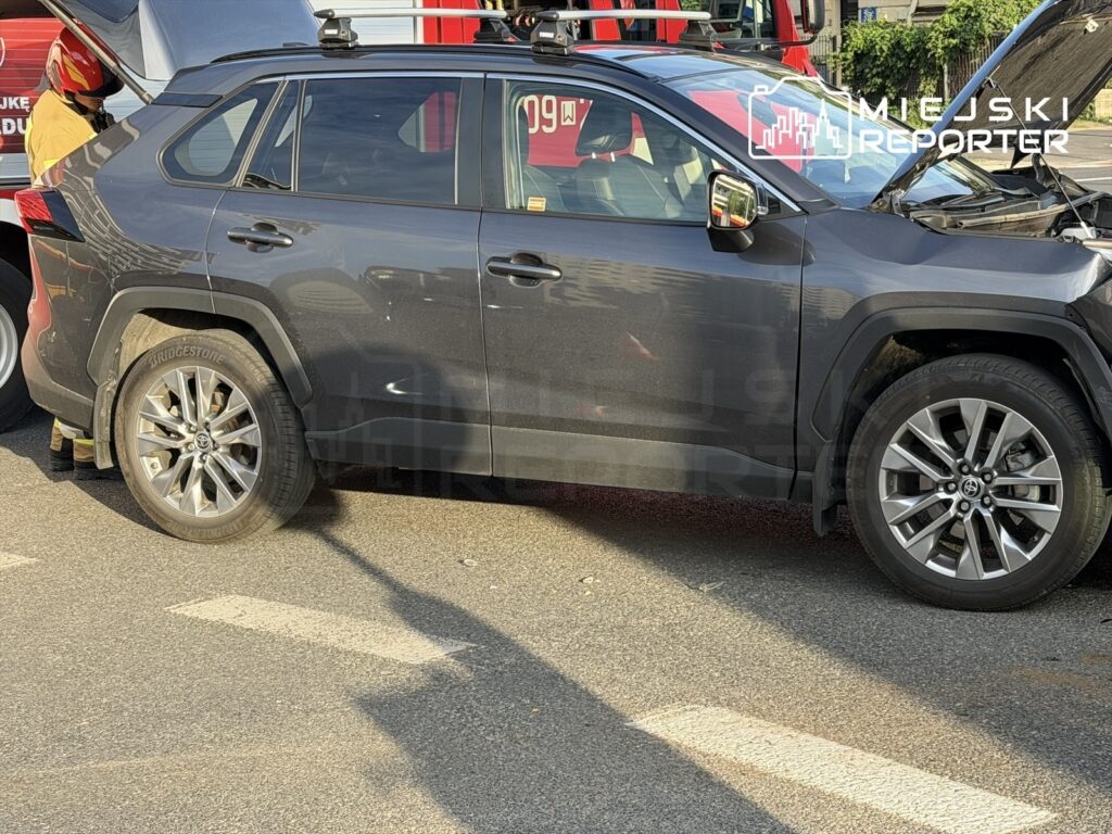 Czarny SUV z otwartą maską stoi na parkingu, obok pojazdu strażackiego z widocznymi reflektorami.