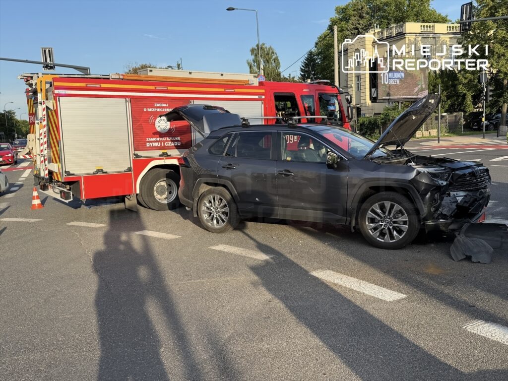 Czarny SUV z uszkodzoną maską stoi obok wozu strażackiego na skrzyżowaniu, otoczony przez inne samochody.