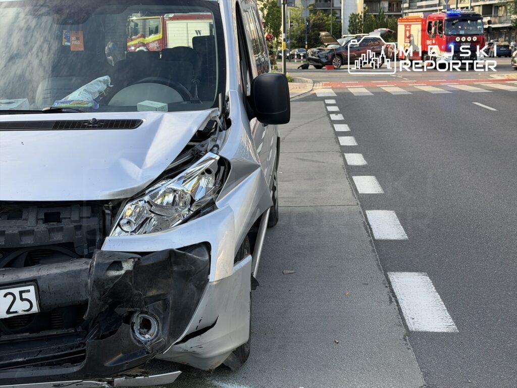Zniszczony przód białego furgonu zaparkowanego na chodniku obok skrzyżowania, w tle widoczny wóz strażacki.