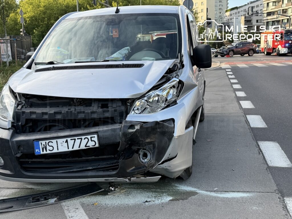 Uszkodzony srebrny samochód dostawczy z wgniecionym przodem stoi na ulicy, obok widoczne inne pojazdy.