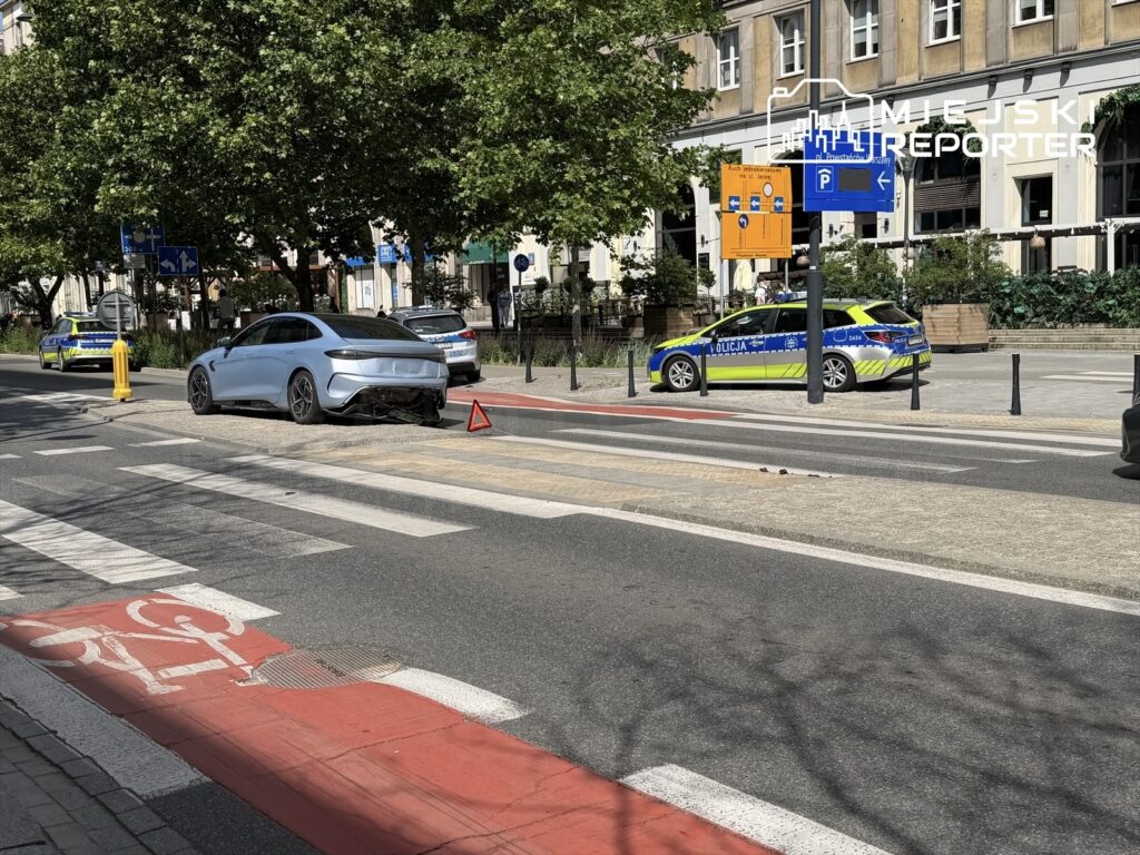 Samochód osobowy z uszkodzeniami stoi na ulicy obok policyjnego radiowozu i oznaczeń drogowych.