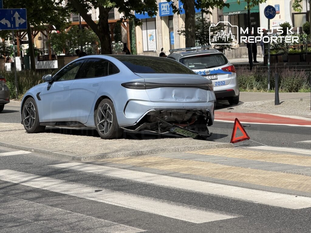 Nieprzygotowany samochód elektryczny w niebieskim kolorze z uszkodzeniem tyłu stoi na ulicy, obok trójkąt ostrzegawczy.
