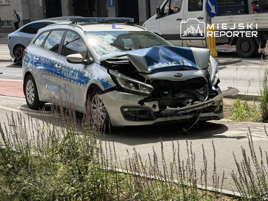 Policyjny samochód osobowy z uszkodzoną przednią częścią stoi na ulicy, obok roślinności.