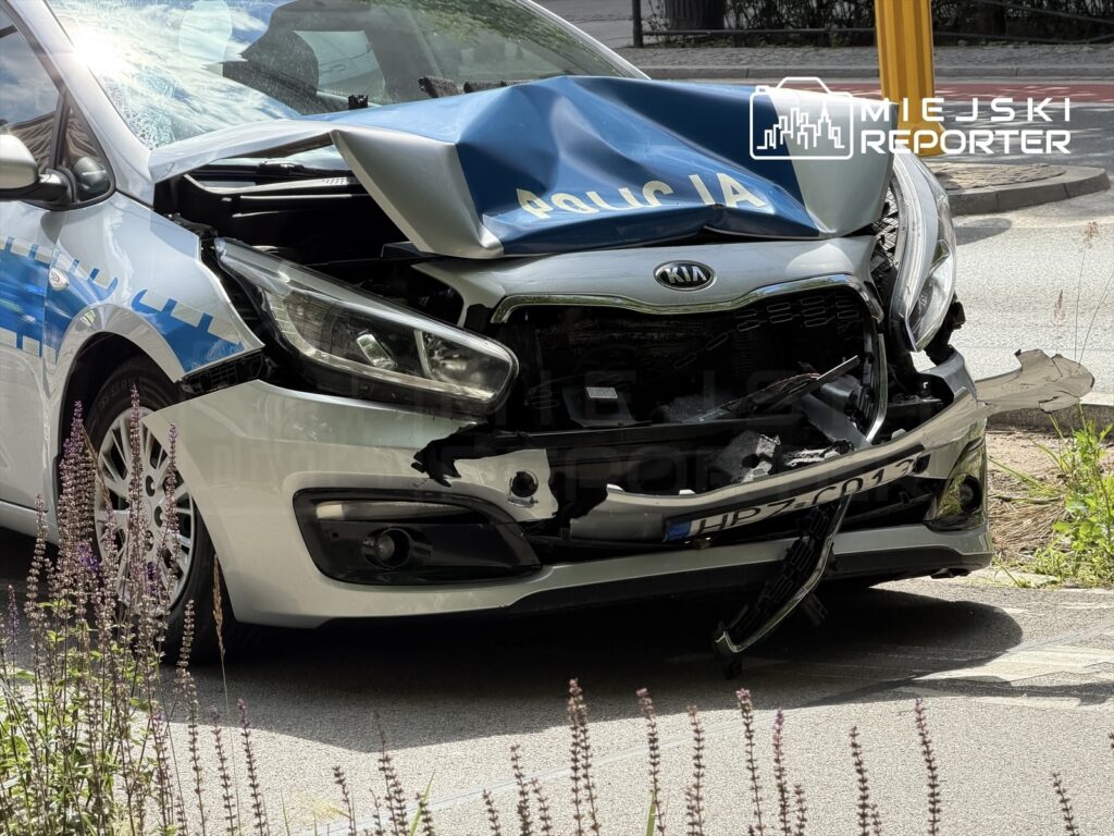 Uszkodzony samochód osobowy marki Kia z wgniecionym przodem stoi na poboczu drogi, otoczony zielenią.
