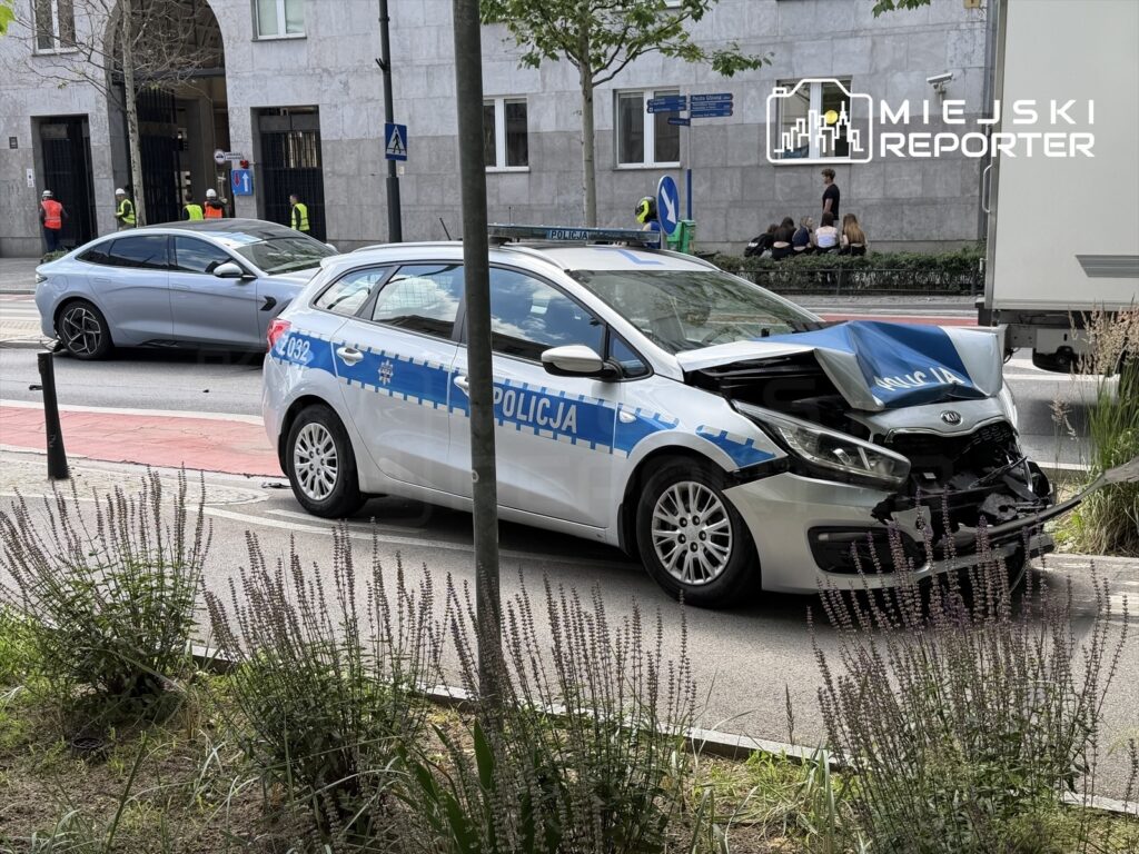 Policyjny samochód z uszkodzoną maską stoi na ulicy, obok widać inny pojazd oraz grupę osób na chodniku.