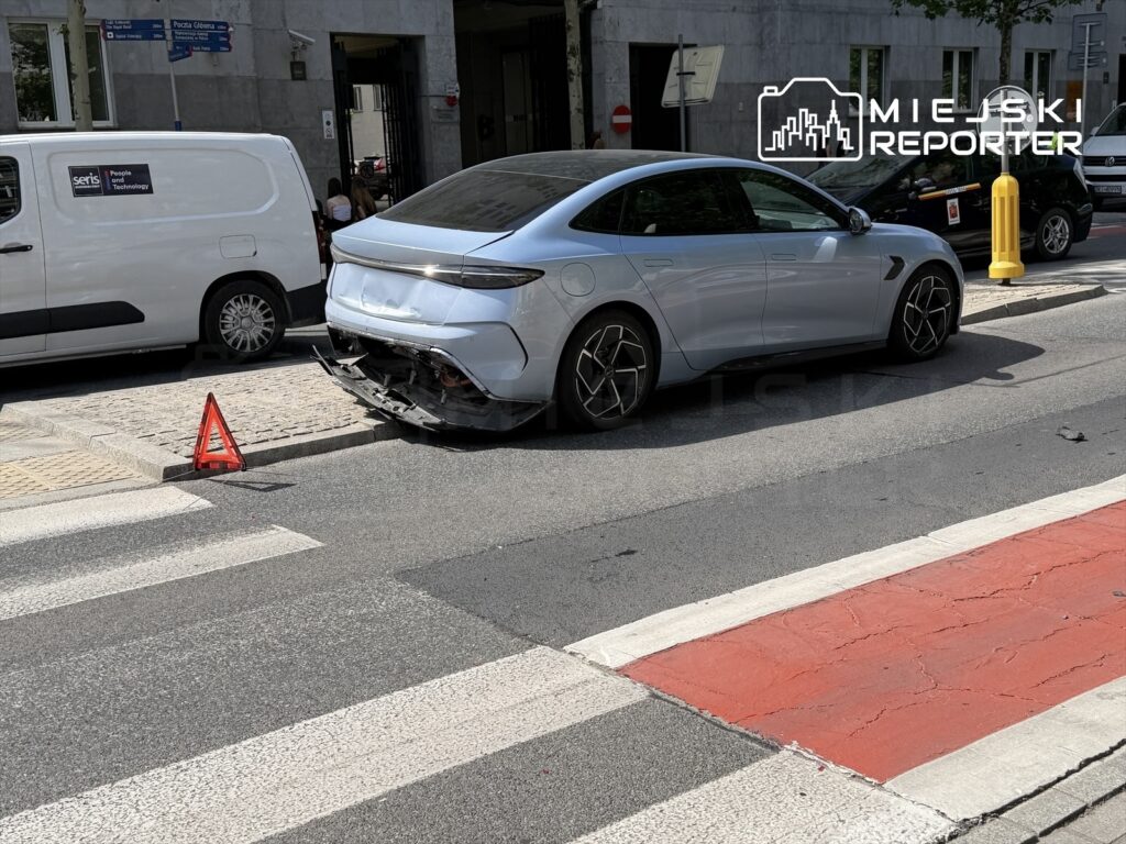 Uszkodzony samochód osobowy z rozbitą tylną częścią zaparkowany na ulicy, obok znajduje się trójkąt ostrzegawczy.