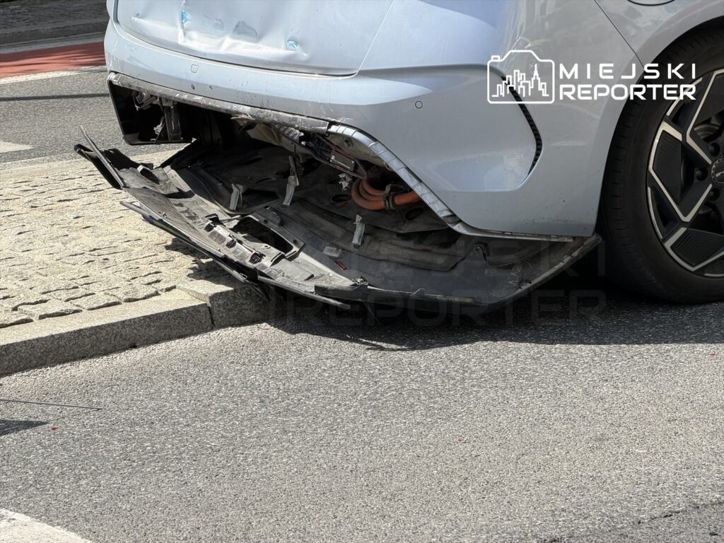 Uszkodzony tył samochodu osobowego z odczepionym zderzakiem na ulicy, obok krawężnika.
