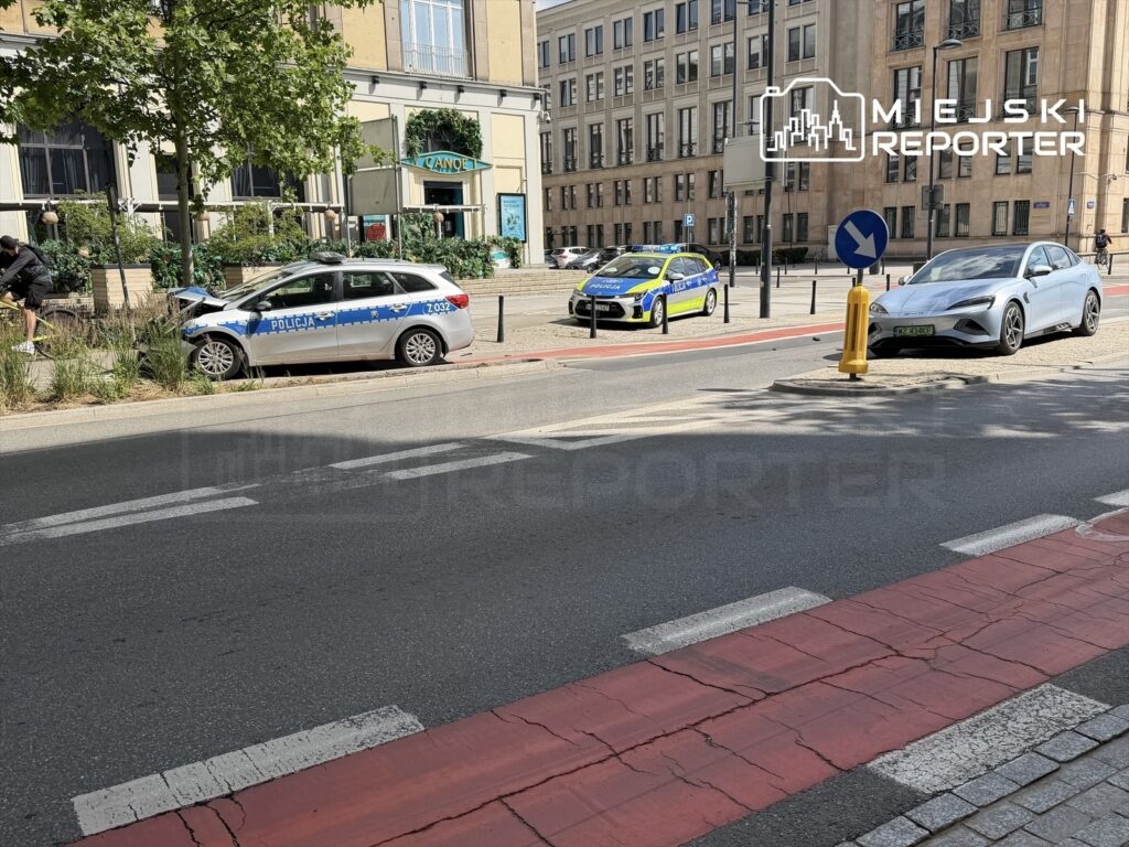 Policyjny radiowóz z uszkodzeniem na parkingu, obok stoi drugi radiowóz, w tle restauracja z zielonymi roślinami.