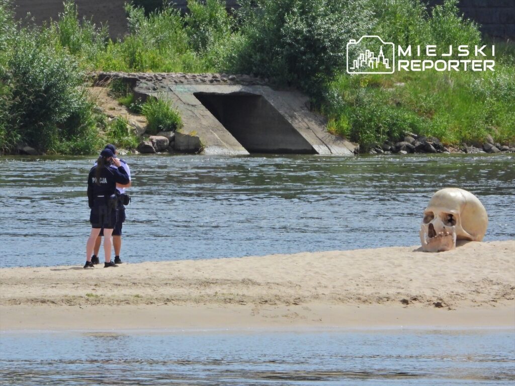 Dwóch policjantów rozmawia na piaszczystym brzegu rzeki, obok dużej czaszki leżącej na plaży.