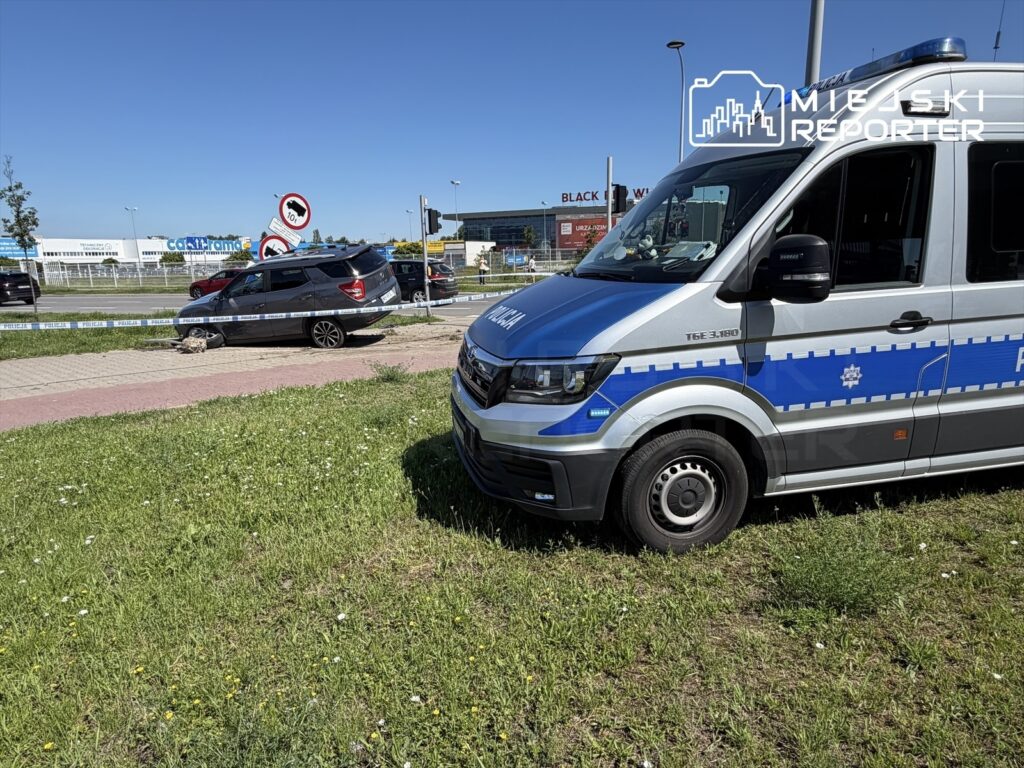 Policyjny radiowóz zaparkowany na trawie obok uszkodzonego samochodu osobowego, otoczony taśmą zabezpieczającą.