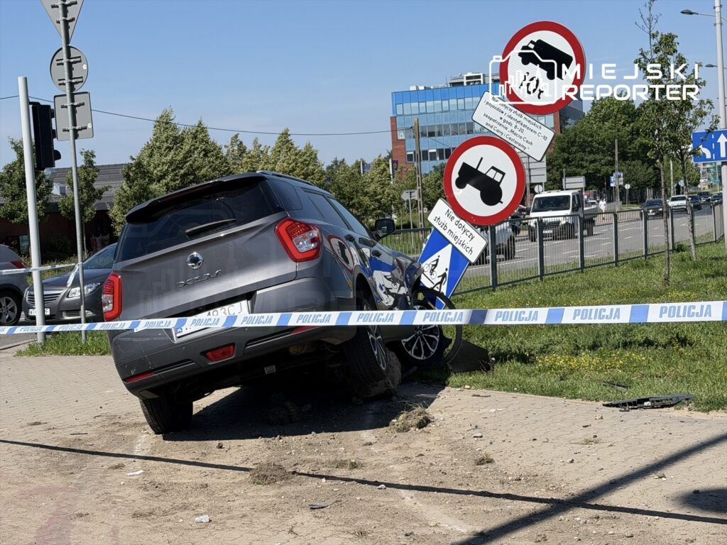 Samochód osobowy stoi na krawężniku, z jedną stroną uniesioną, obok znaków drogowych z ostrzeżeniem.