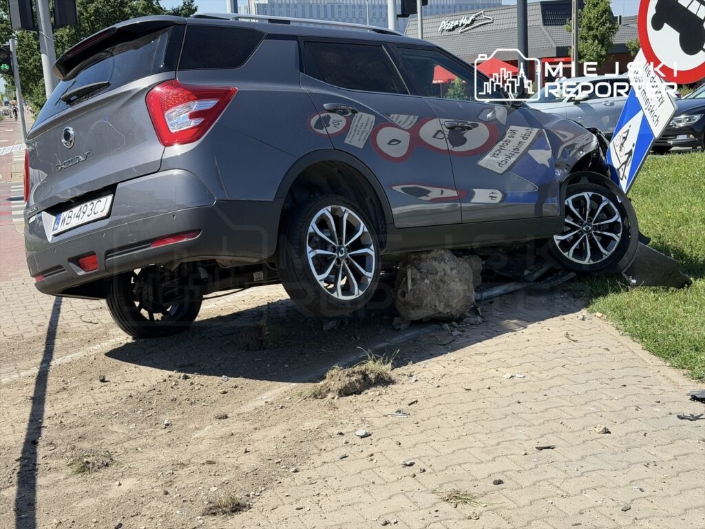 Samochód osobowy unosi się na krawężniku, opierając się na głazie, obok znak drogowy.