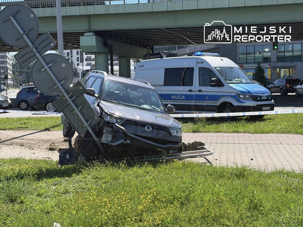 Czarny SUV wjeżdża w znak drogowy na trawniku, obok stoi policyjny radiowóz z włączonymi sygnałami.