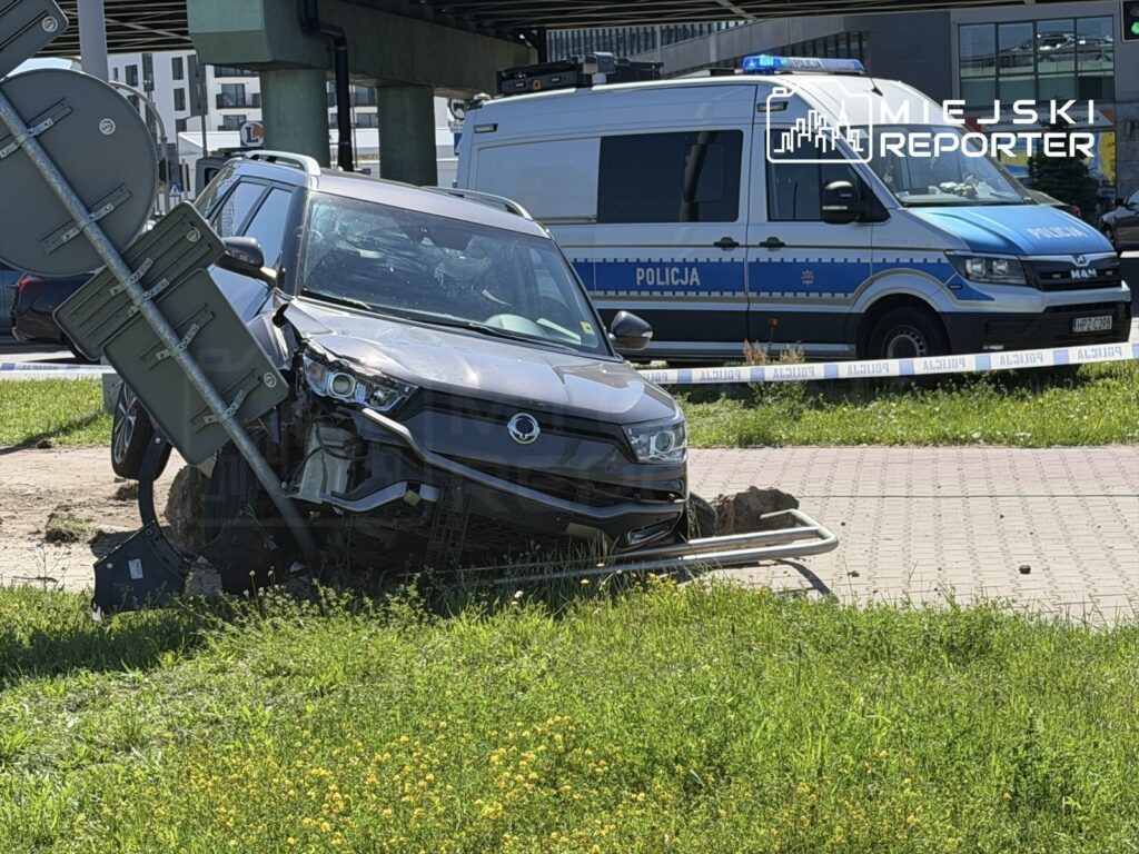 Uszkodzony samochód osobowy stoi na trawie obok słupa z tablicą drogową, a w tle widać radiowóz policji.
