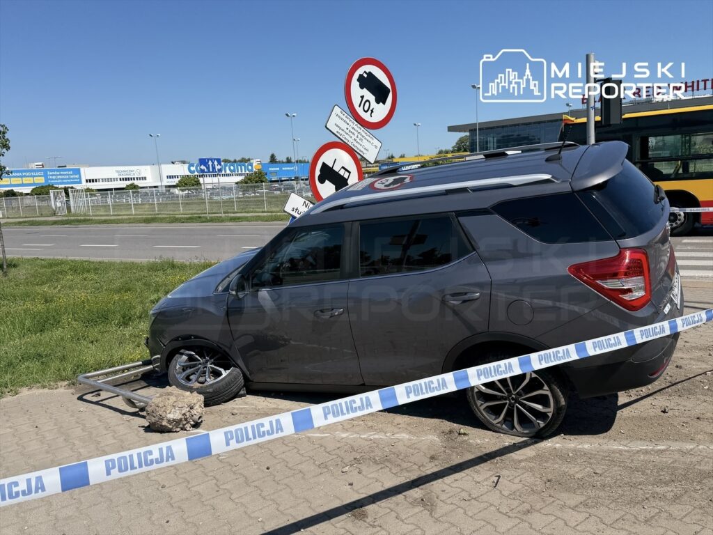 Samochód osobowy stoi na poboczu, z uszkodzoną oponą, otoczony taśmą policyjną. W tle widoczne znaki drogowe i budynki.