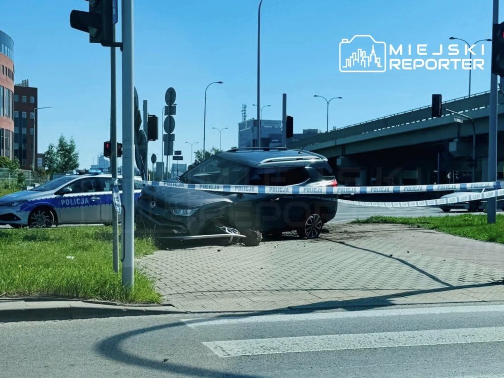 Czarny samochód osobowy z uszkodzoną przednią częścią stoi na trawniku, otoczony taśmą policyjną. W tle widoczny radiowóz.