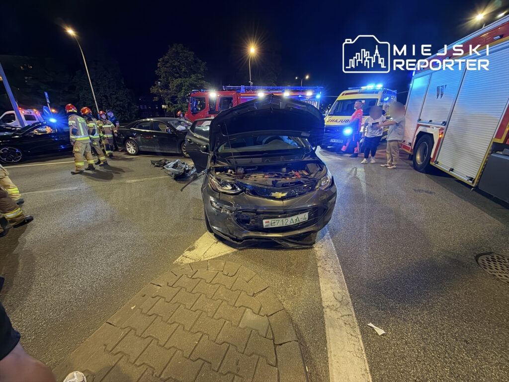 Na skrzyżowaniu po nocnym wypadku drogowy, strażacy udzielają pomocy poszkodowanym, obok uszkodzonego samochodu.