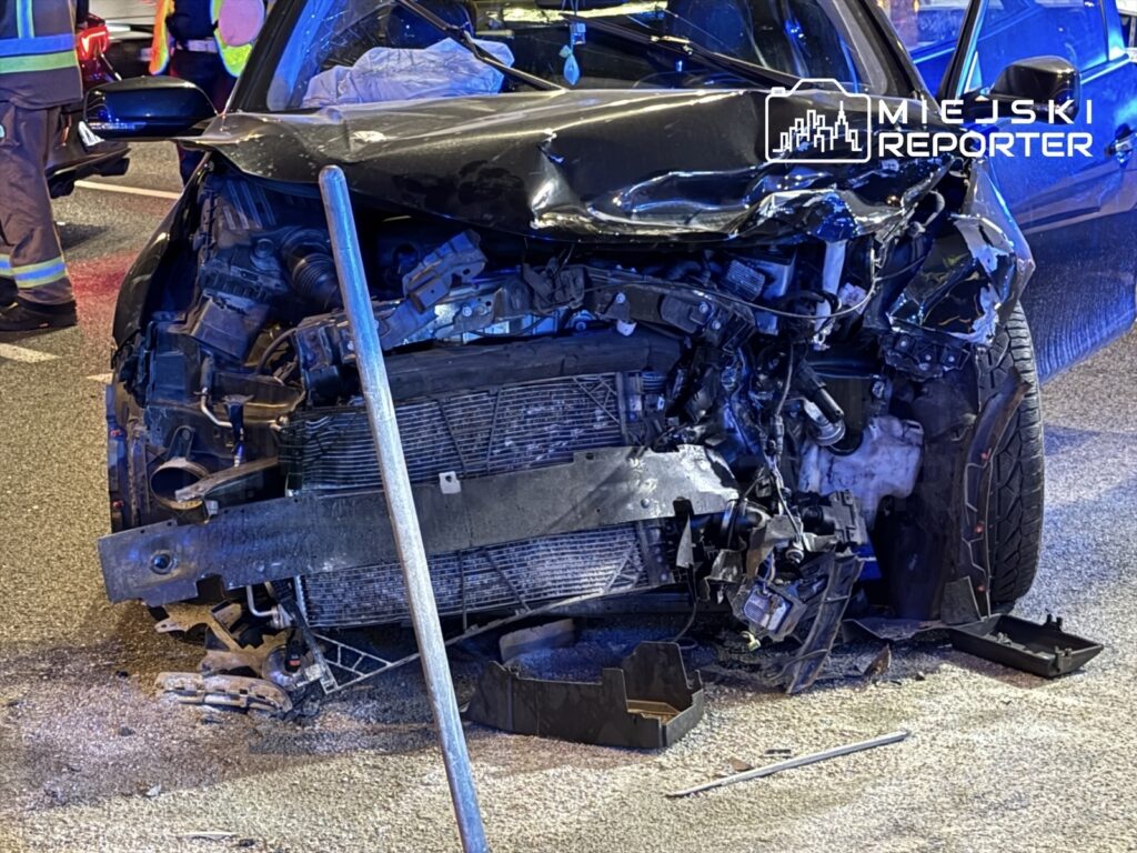 Zniszczony przód czarnego samochodu osobowego na drodze, z widocznymi uszkodzeniami i fragmentami pojazdu na jezdni.