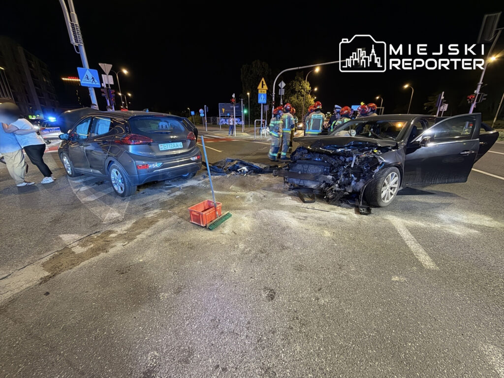 Dwa uszkodzone samochody po kolizji na ulicy, w tle strażacy i osoby zabezpieczające miejsce zdarzenia.
