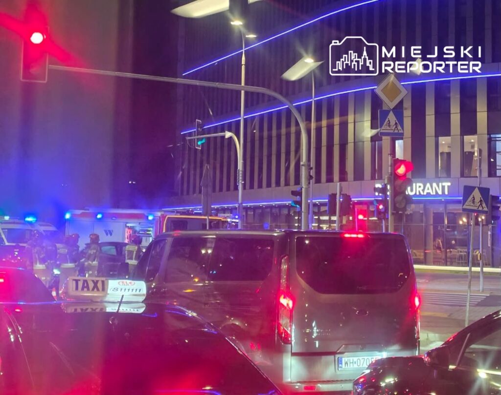W nocy, w ruchliwej okolicy, samochody stoją w korku, a w tle widać pojazdy służb ratunkowych oraz policję.