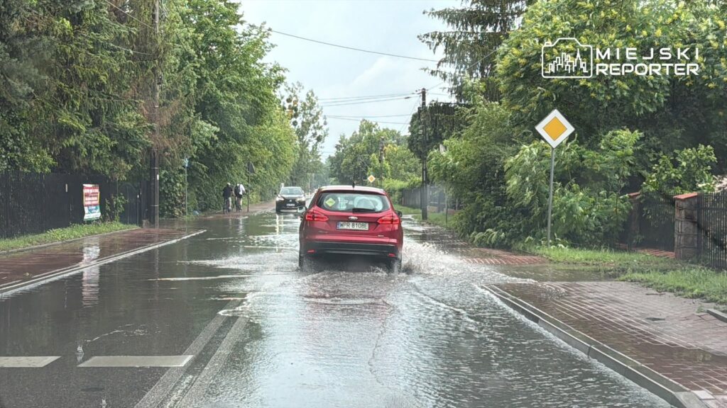 Czerwony samochód osobowy jedzie przez zalaną ulicę, obok spacerują piesi i rowerzyści w deszczowej aurze.