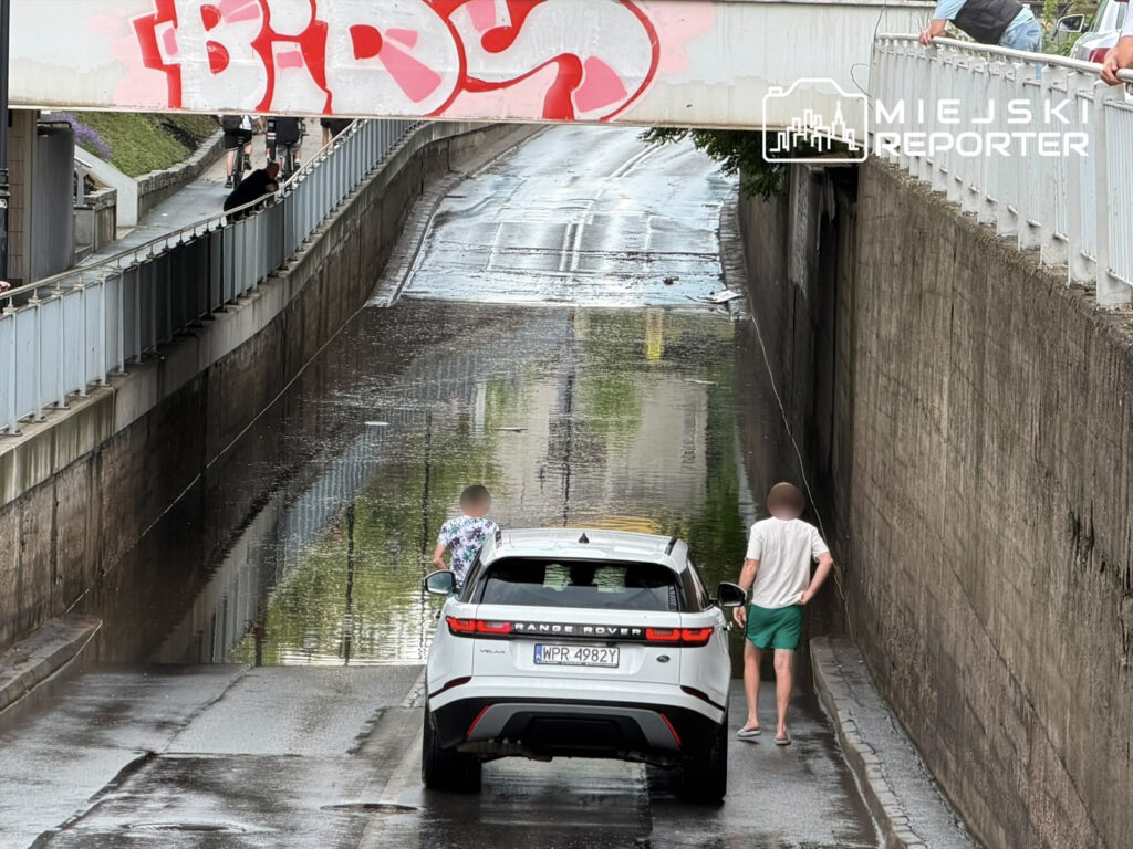 Biały samochód terenowy stoi na mokrej drodze, obok dwie osoby spacerują wzdłuż kanału, a nad nimi widoczny graffiti.