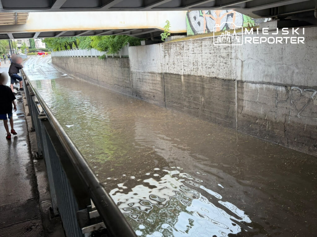 Dzieci spacerujące wzdłuż zalanego chodnika pod mostem, z widoczną wodą odbijającą otoczenie.