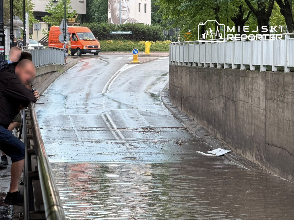 Grupa mężczyzn opiera się o barierkę, obserwując zalaną wodą ulicę po deszczu. W tle widoczny pomarańczowy samochód dostawczy.