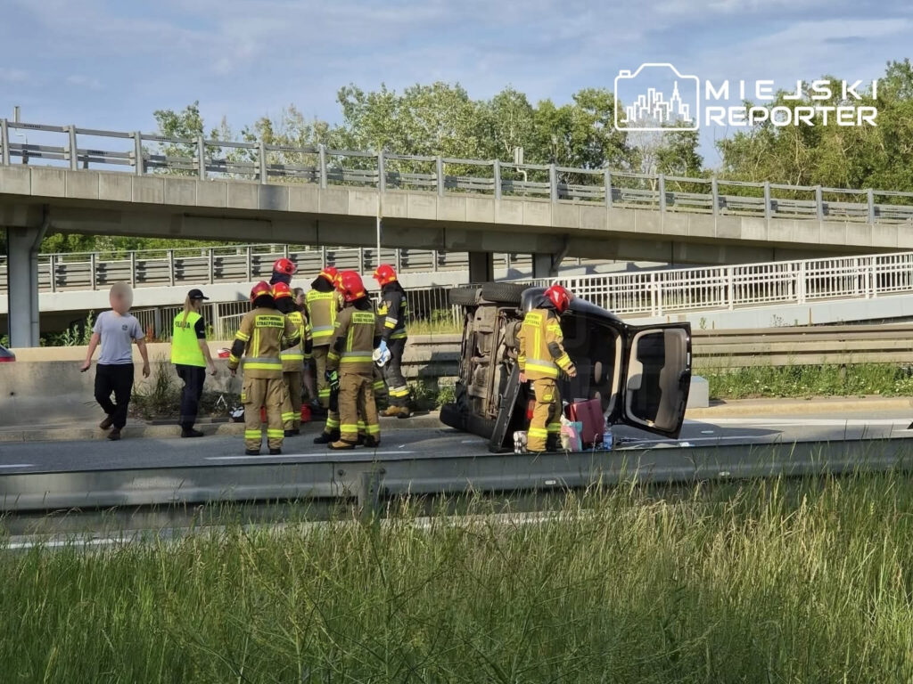 Grupa strażaków w mundurach i kaskach pracuje przy przewróconym samochodzie na drodze, obok nich policjant rozmawia z osobą.