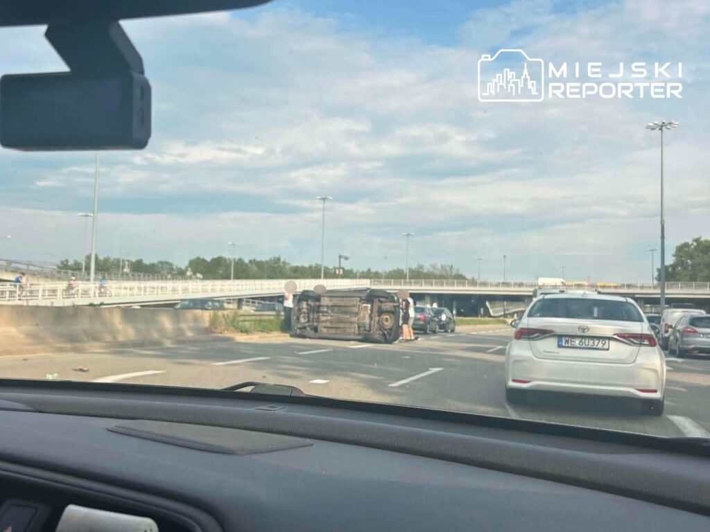 Na drodze leży przewrócone auto, a obok zatrzymali się kierowcy, obserwując sytuację. W tle widoczny most.