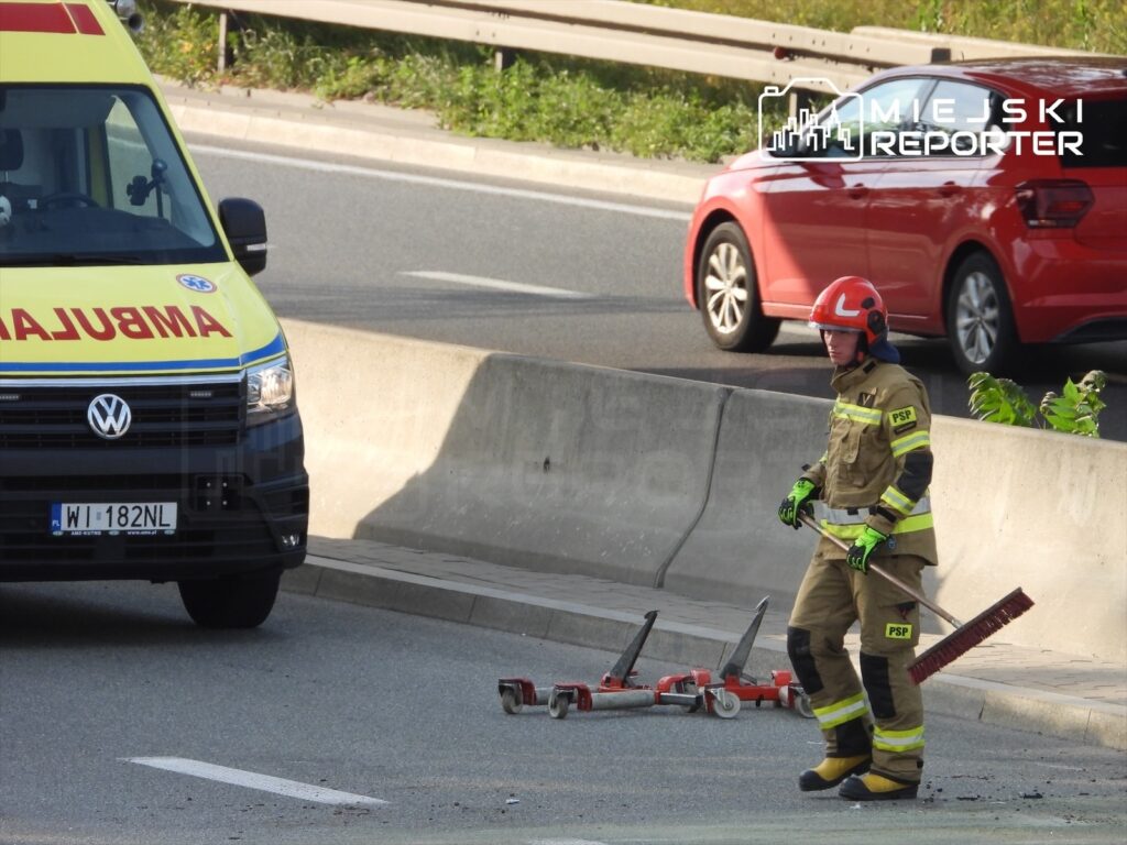Strażak w kombinezonie ochronnym sprząta pozostałości po wypadku na drodze, obok stoi ambulans.