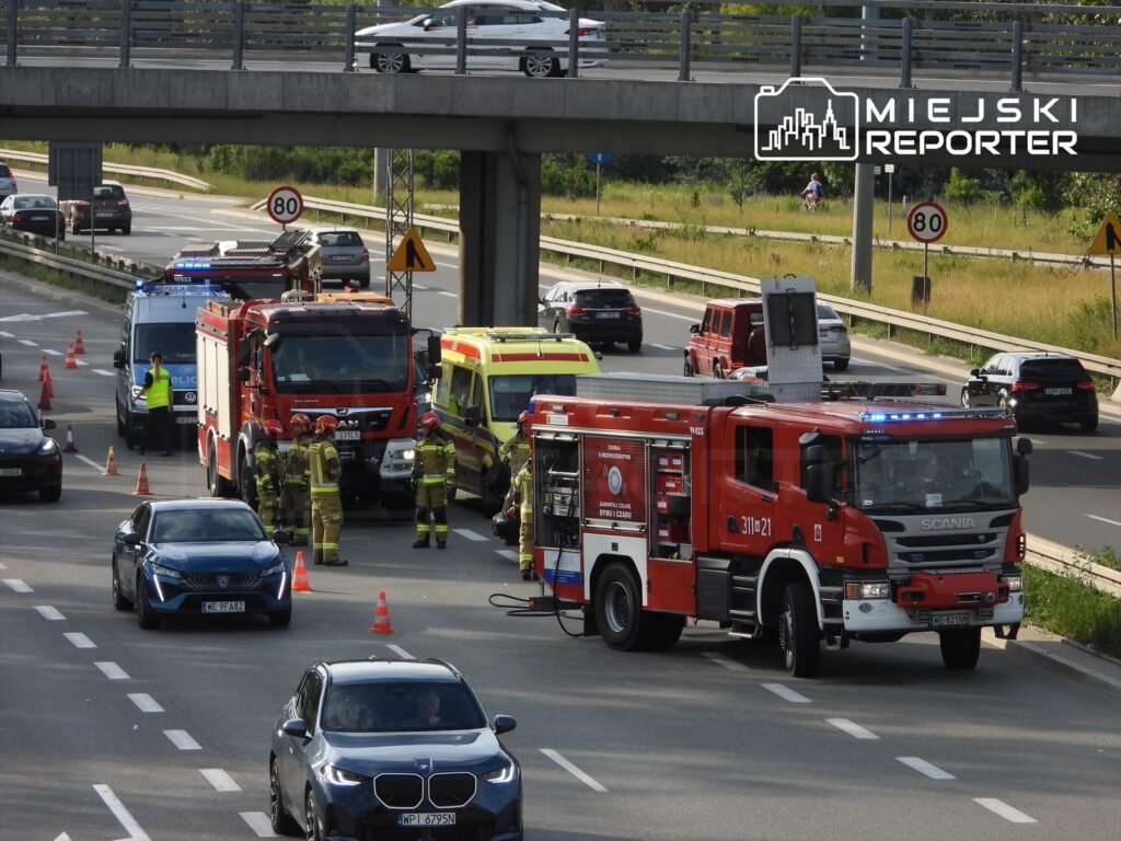 Na drodze zatrzymane pojazdy, w tym wóz strażacki i karetka, a strażacy oraz policjanci prowadzą działania ratunkowe.