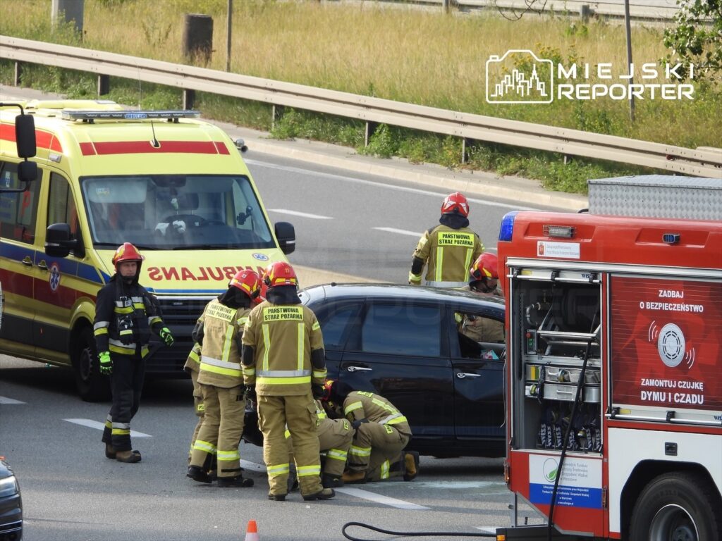 Grupa strażaków w mundurach pracuje przy czarnym samochodzie na drodze, obok stoi karetka i wóz strażacki.