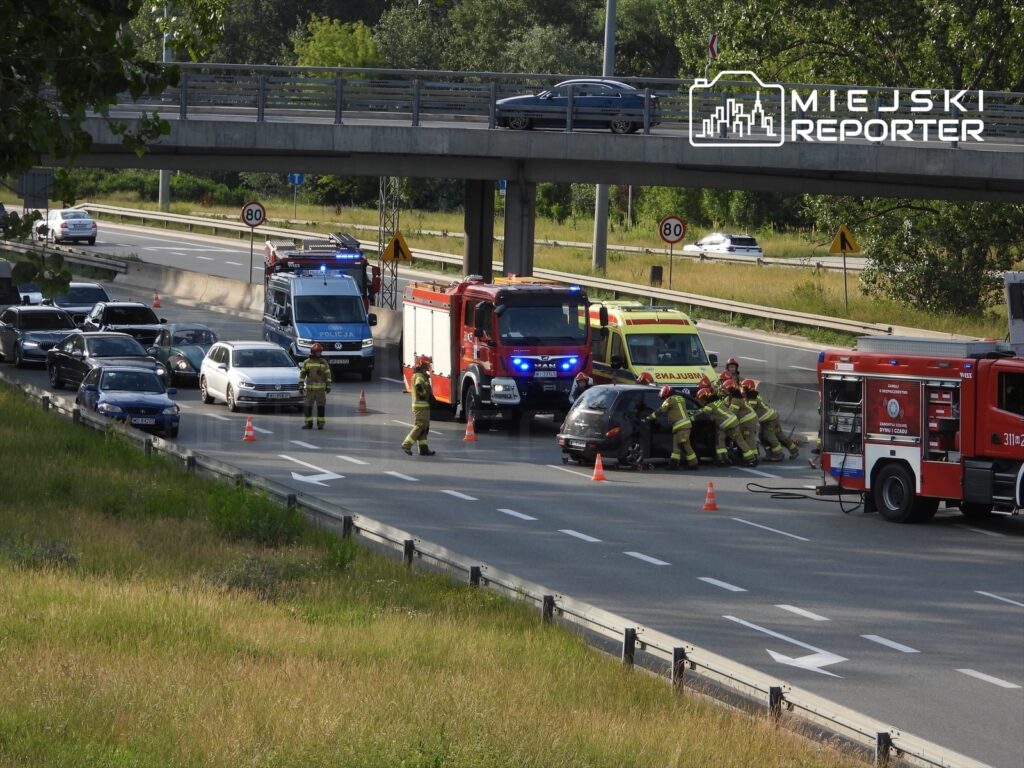 Grupa strażaków udziela pomocy przy wypadku drogowym na jezdni, obok zaparkowanych pojazdów.