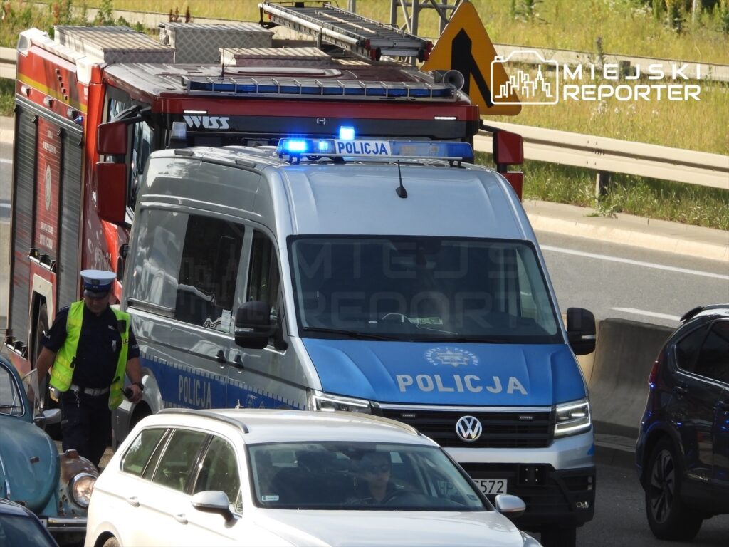 Policjant w kamizelce odblaskowej kontroluje ruch obok radiowozu i wozu strażackiego na zablokowanej drodze.