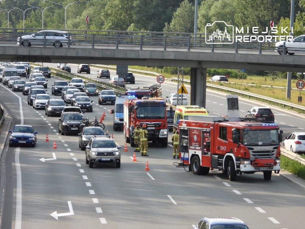 Ruch samochodowy zatrzymany na drodze, w tle straż pożarna i karetka, w pobliżu wypadku. Czerwone pachołki ostrzegawcze ustawione wzdłuż jezdni.