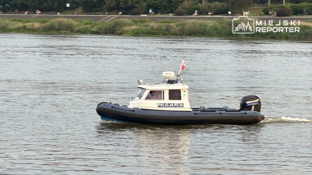 Policyjny kuter motorowy pływający po rzece, z flagą i oznaczeniem "POLICJA PZ 36". W tle widać zieleń i ścieżkę rowerową.