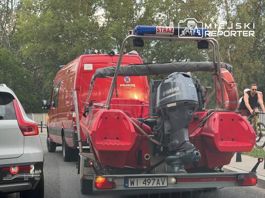 Czerwony pojazd straży pożarnej z przyczepioną łodzią motorową stoi na drodze, obok przejeżdża rowerzysta.