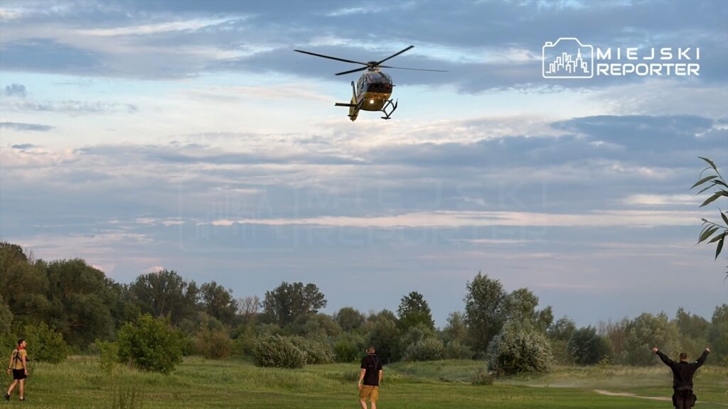 Helikopter przelatujący nad zielonym terenem, w tle drzewa i chmury. Dwie osoby machają rękami w kierunku maszyny.