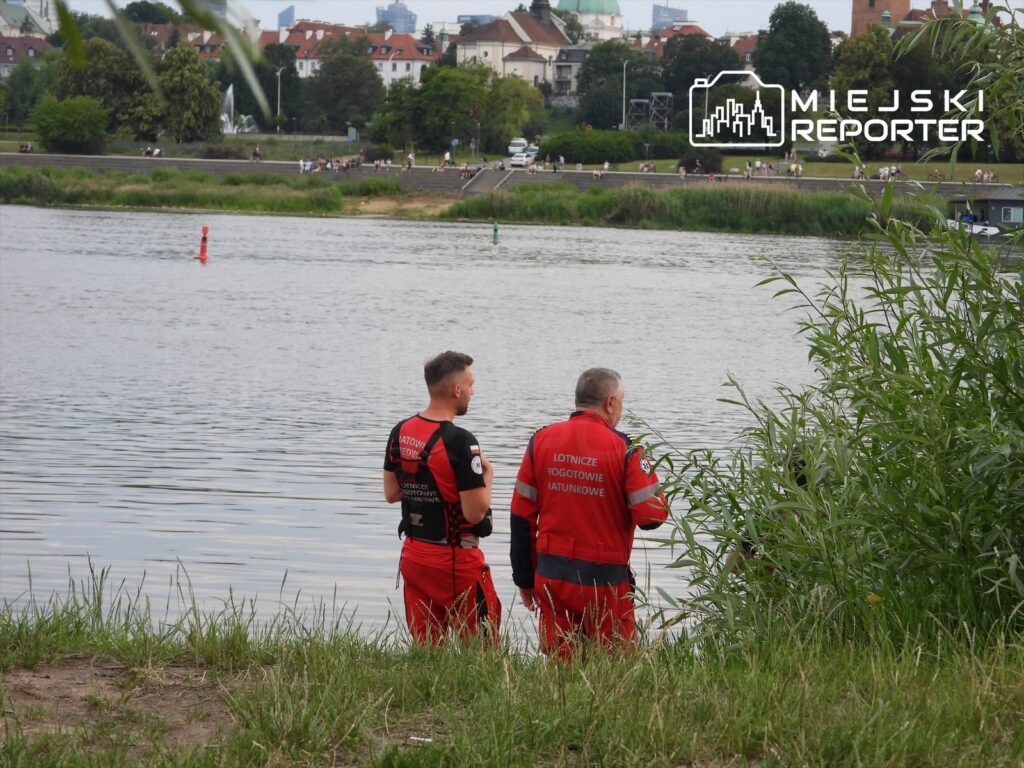 Dwaj ratownicy w czerwonych strojach stoją nad rzeką, obserwując wodę w towarzystwie zieleni.